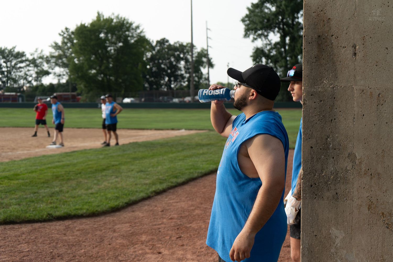 Man in blue sleeveless shirt drinks water bottle leaning against wall at outdoor baseball field.