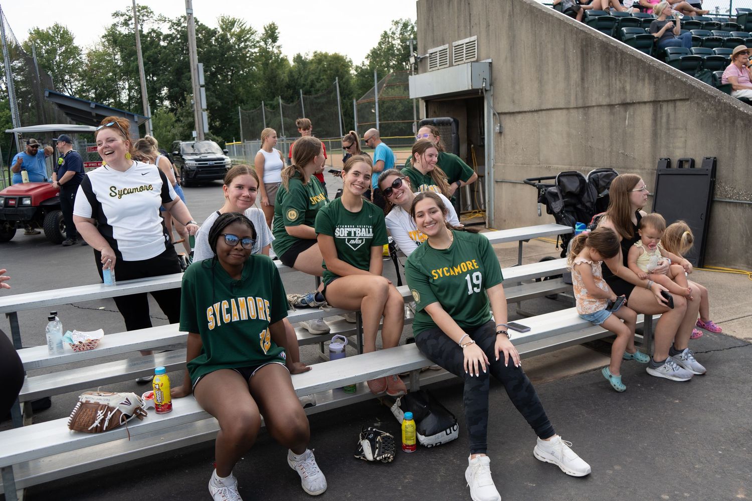 Sycamore team girls sitting on bleachers in green and gold uniforms at an outdoor sports event.