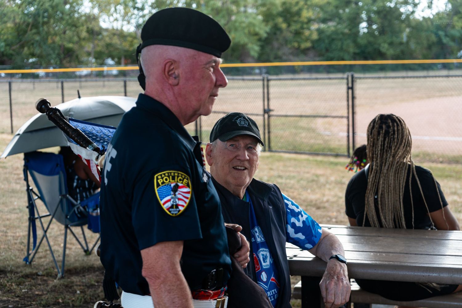Two older men in police uniforms sitting at a picnic table outdoors at a baseball field.