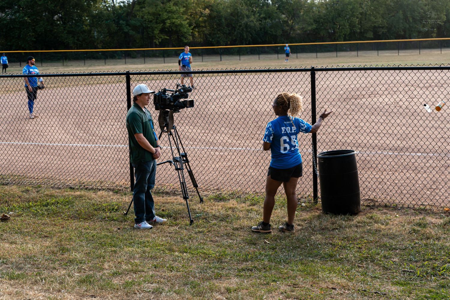 Cameraman filming a player near a chain-link fence at an outdoor softball field.