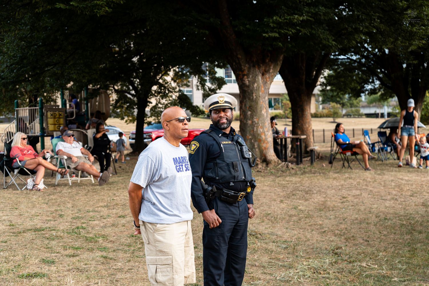 A civilian in a white shirt stands beside a uniformed police officer at an outdoor community event.