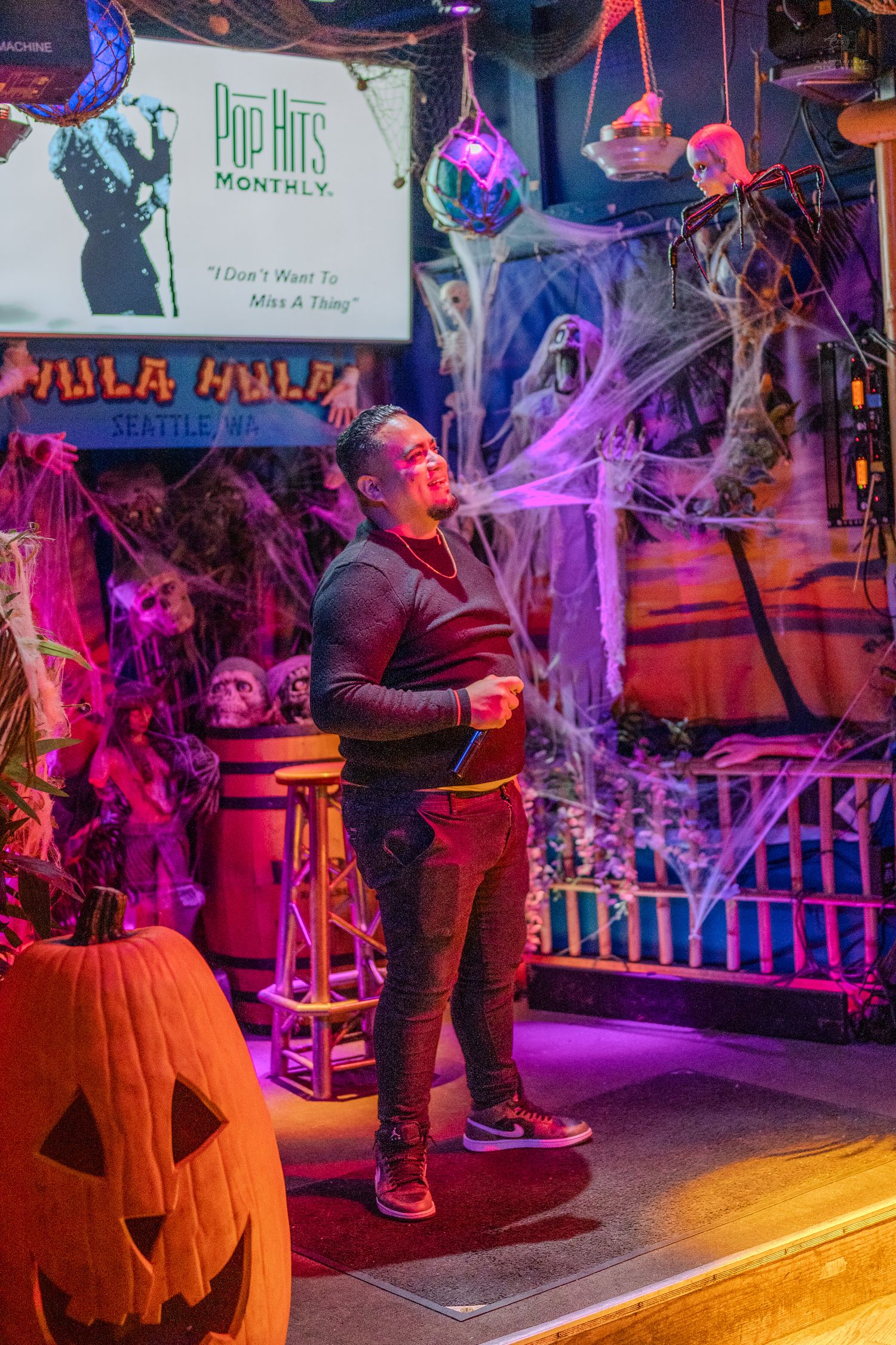 Man performing karaoke at Hula Hula bar in Seattle, surrounded by Halloween decorations and a jack-o-lantern.