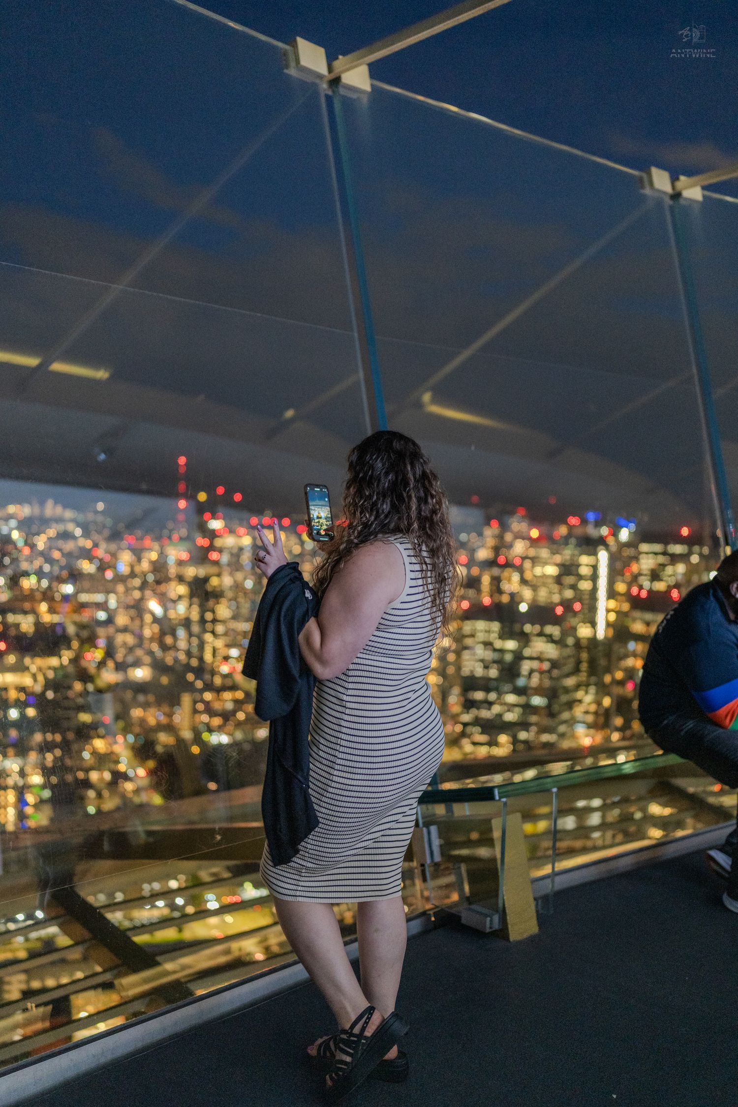 A woman in a striped dress photographs a vibrant city skyline from an observation deck at night.