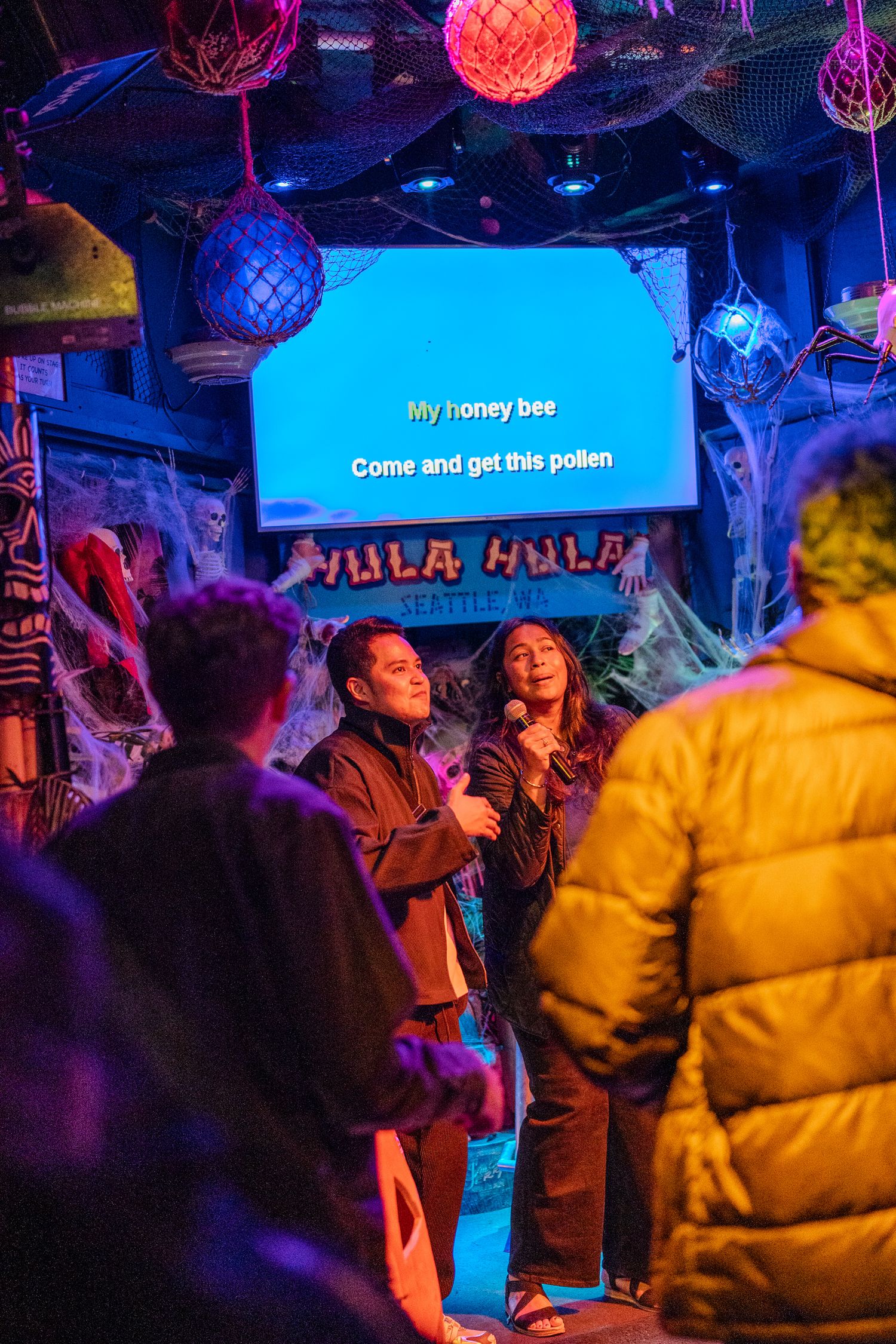 Two friends singing karaoke duet at Hula Hula Seattle bar with festive Halloween and tropical decorations.