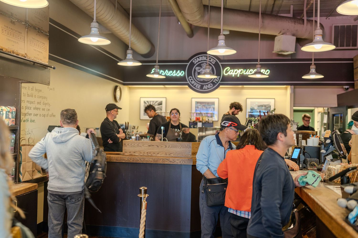 Busy coffee shop interior with baristas serving customers at a wooden counter under pendant lights and espresso signs.