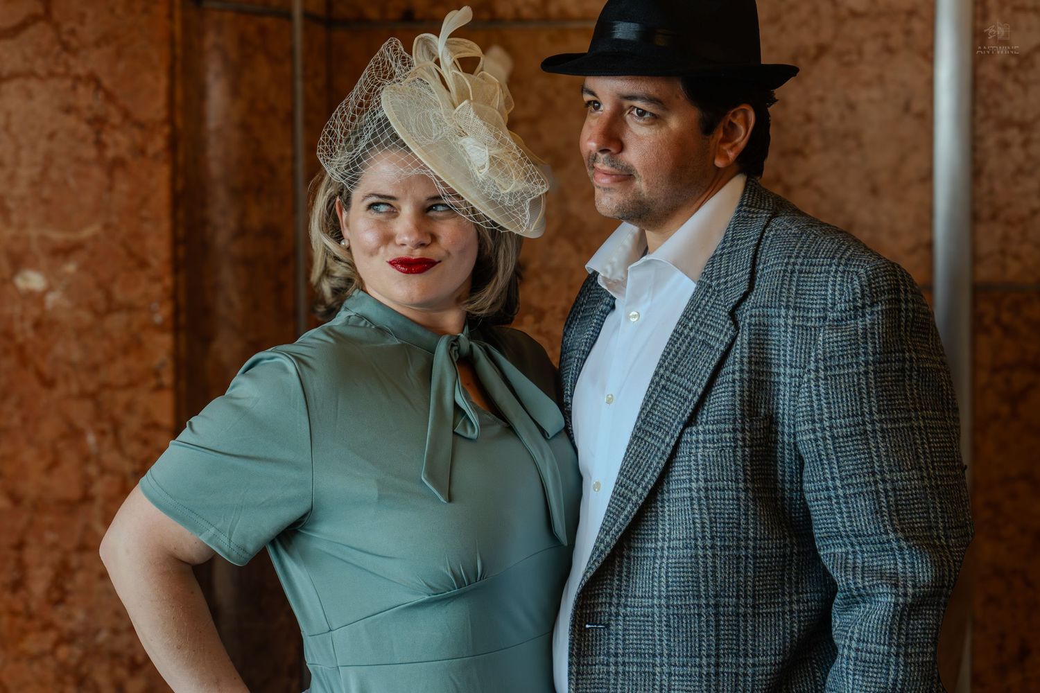 Couple in vintage-inspired outfits, woman in sage green dress with fascinator, man in plaid blazer and black fedora.
