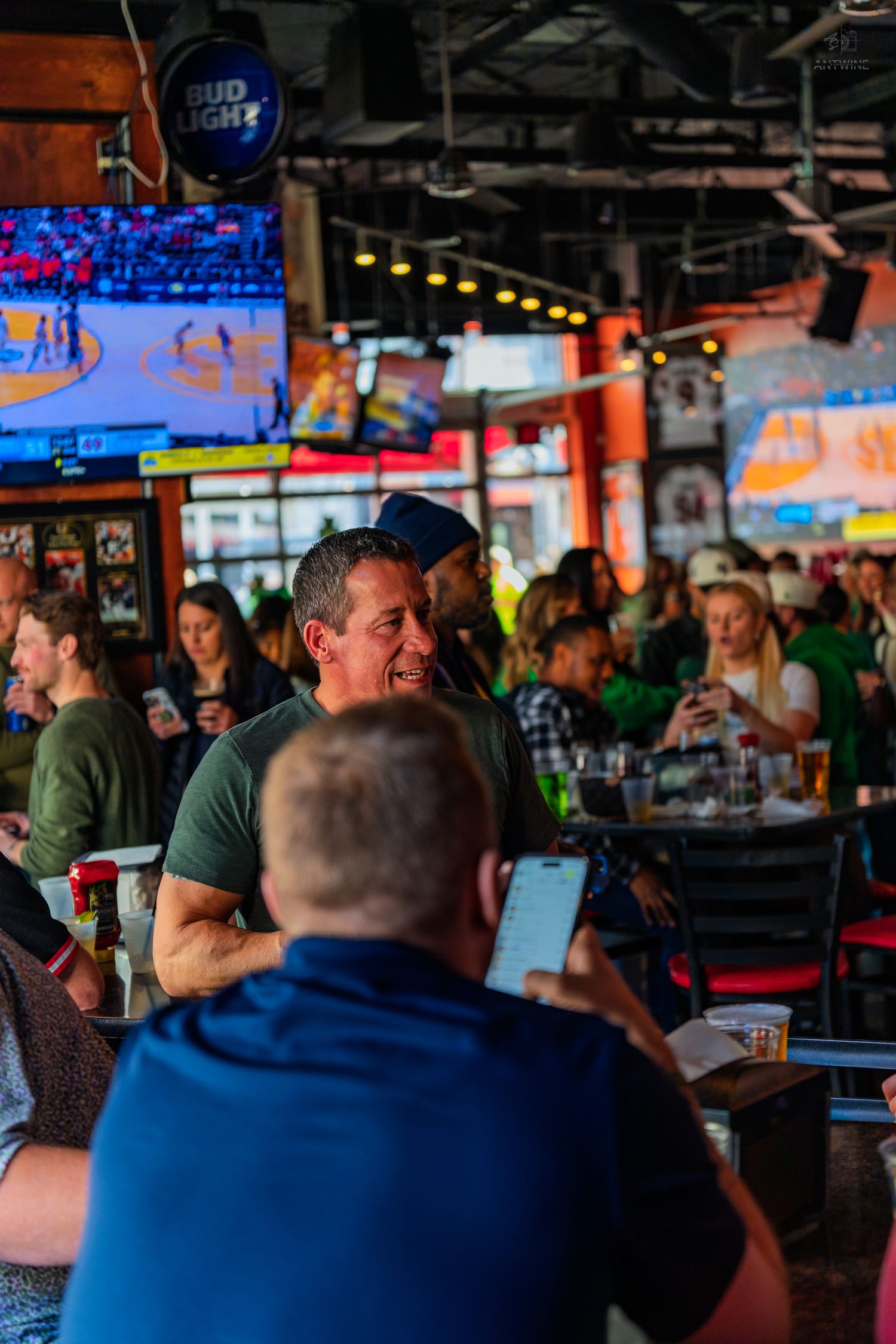 Crowded sports bar with Bud Light sign, TVs showing basketball, and patrons socializing in a lively atmosphere.