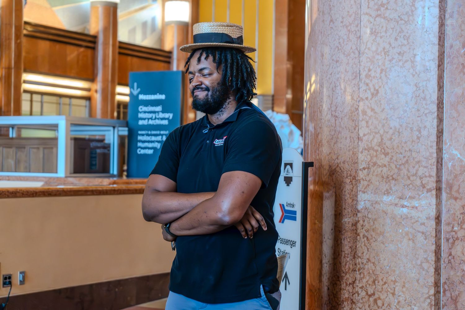 Man with dreadlocks and hat stands arms crossed in a library lobby near an information sign.