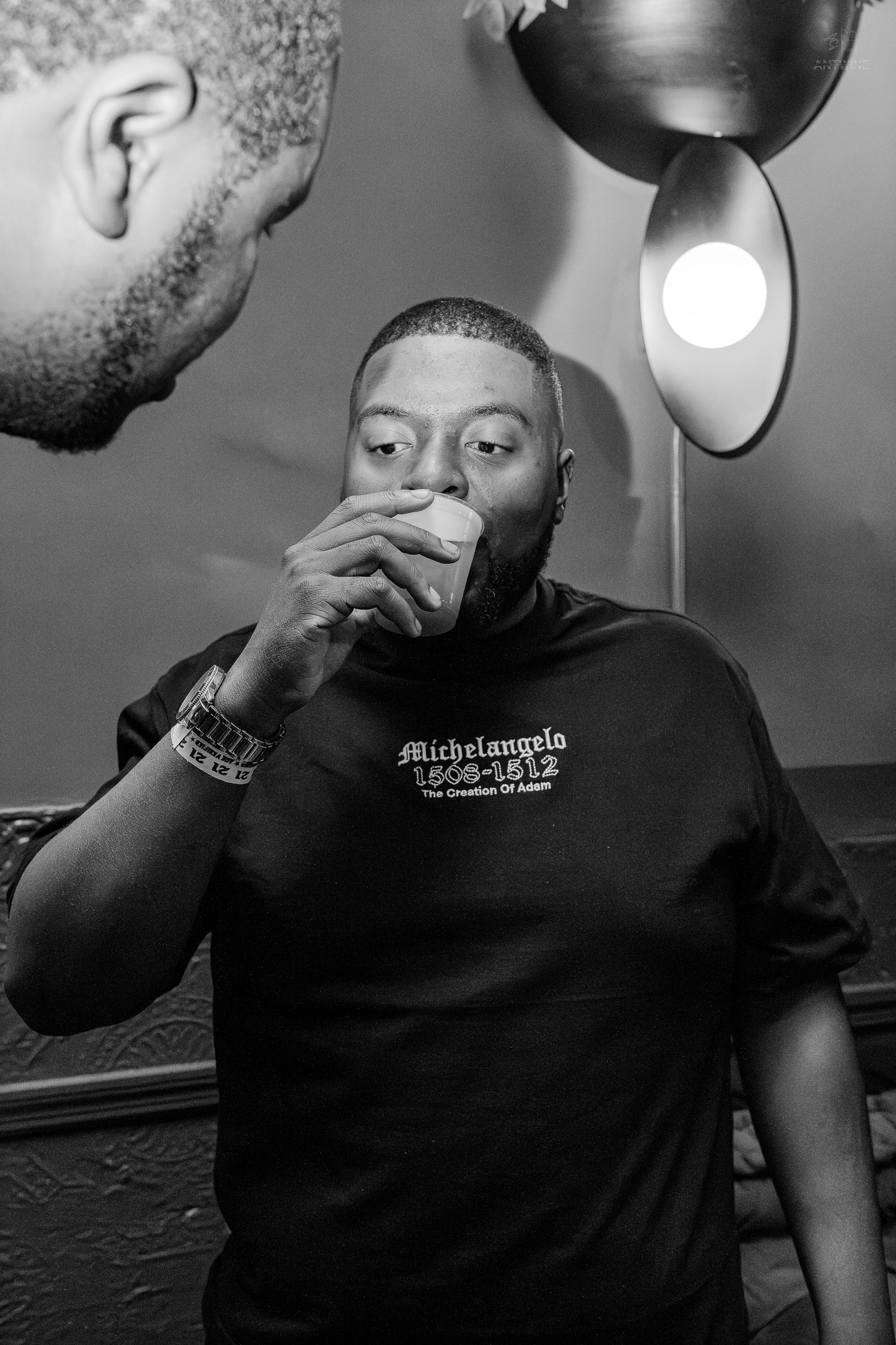 Black and white photo of a man drinking from a cup wearing a Michelangelo t-shirt indoors.