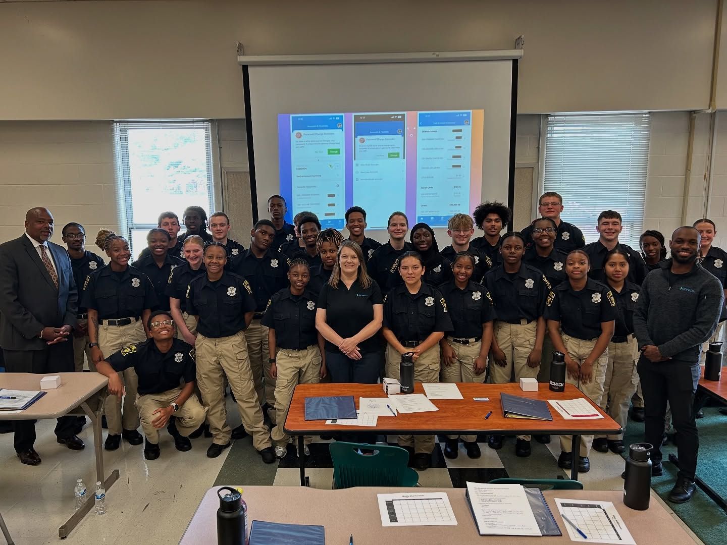 Large group of students in matching uniforms posing for a photo in a classroom with a projector screen.