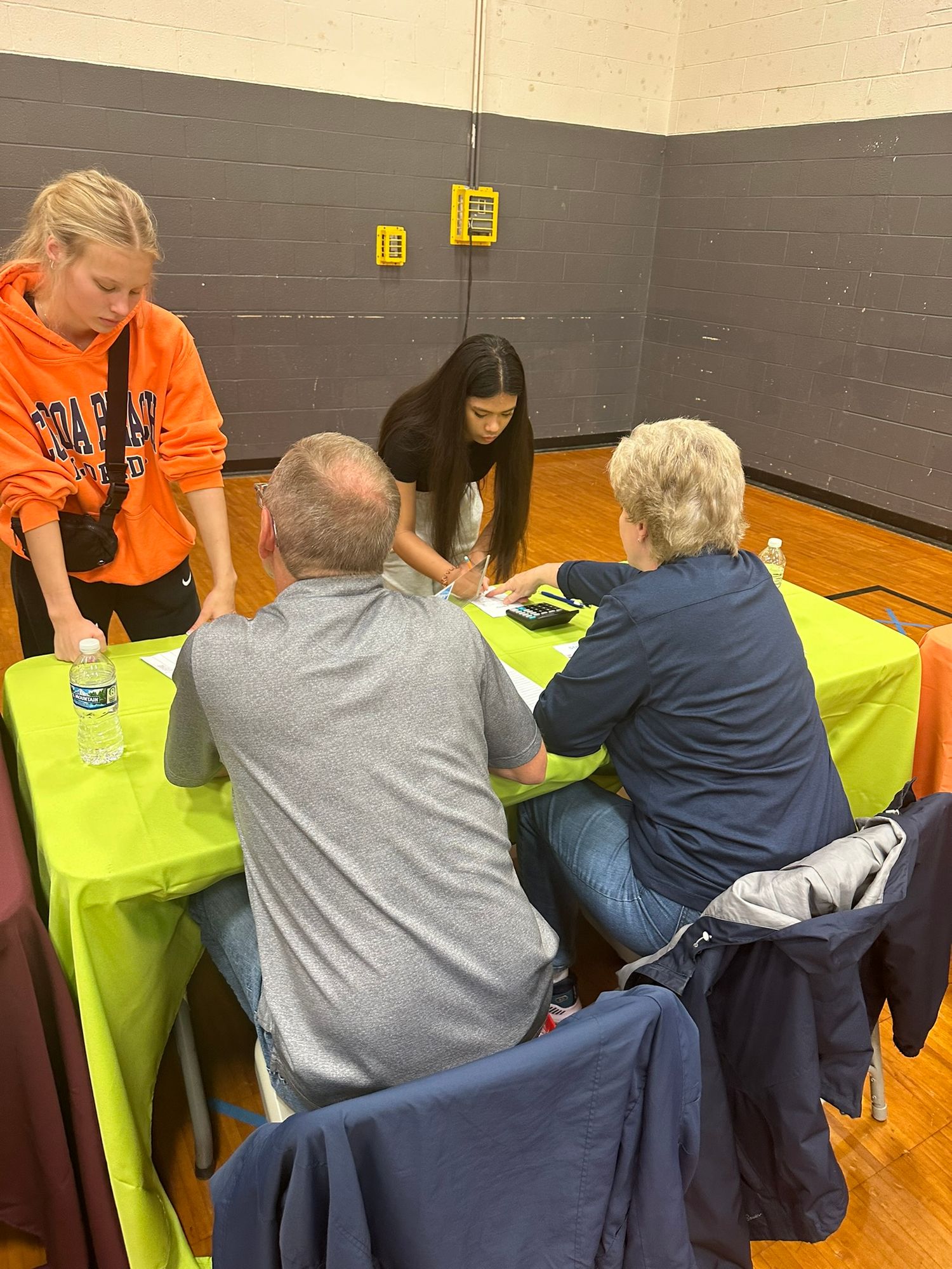 People gathered around a green table in a gymnasium, reviewing documents during a community meeting or health event.