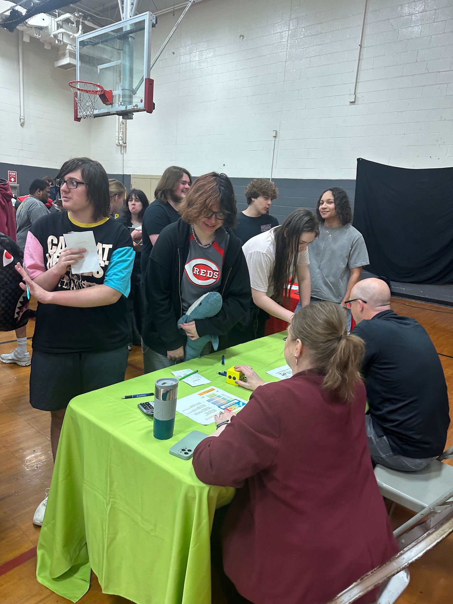 Students gather around a green-draped table in a school gymnasium during a career fair event.