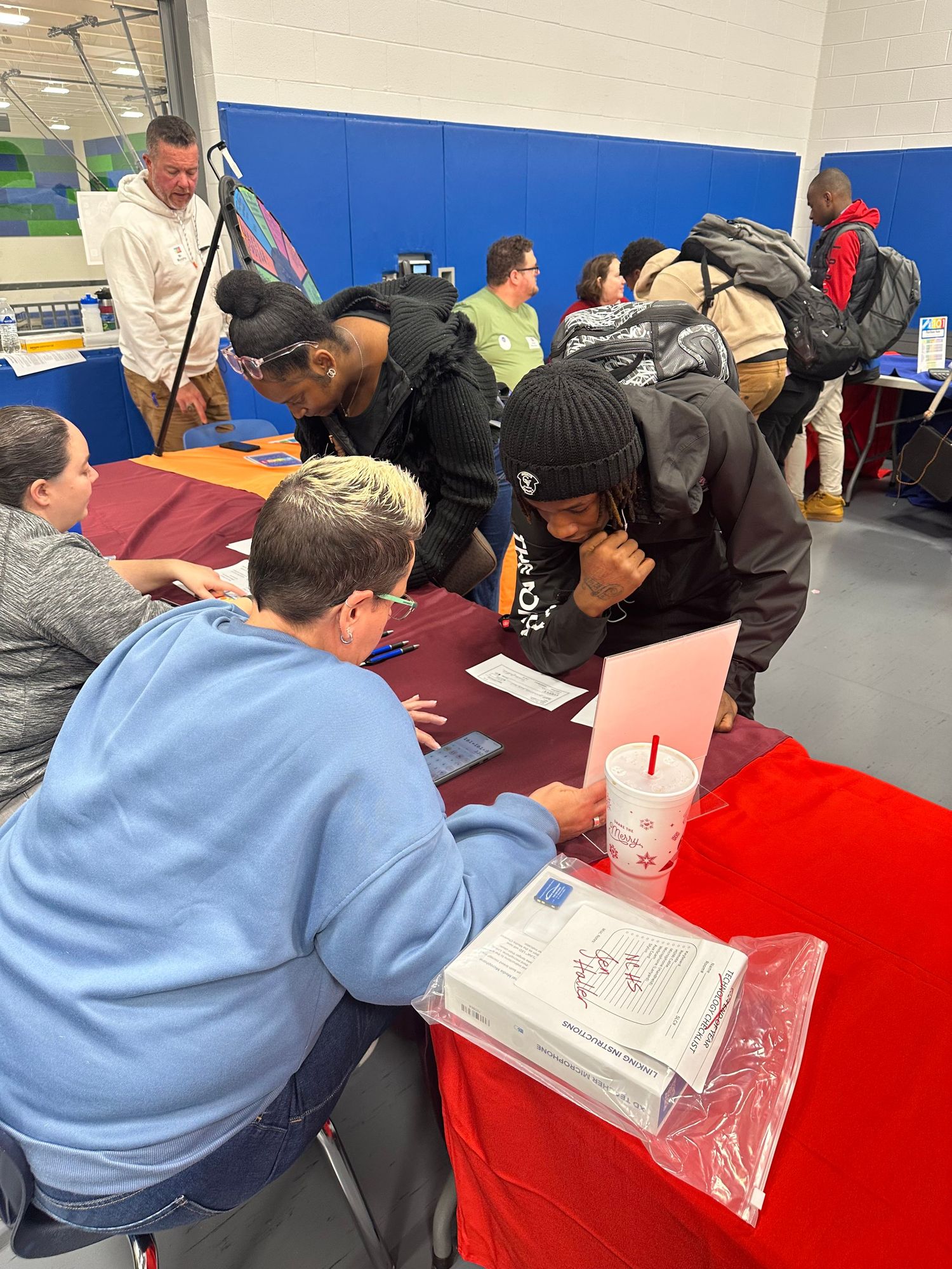 Students fill out forms at a red table during a school career fair event in a gymnasium.