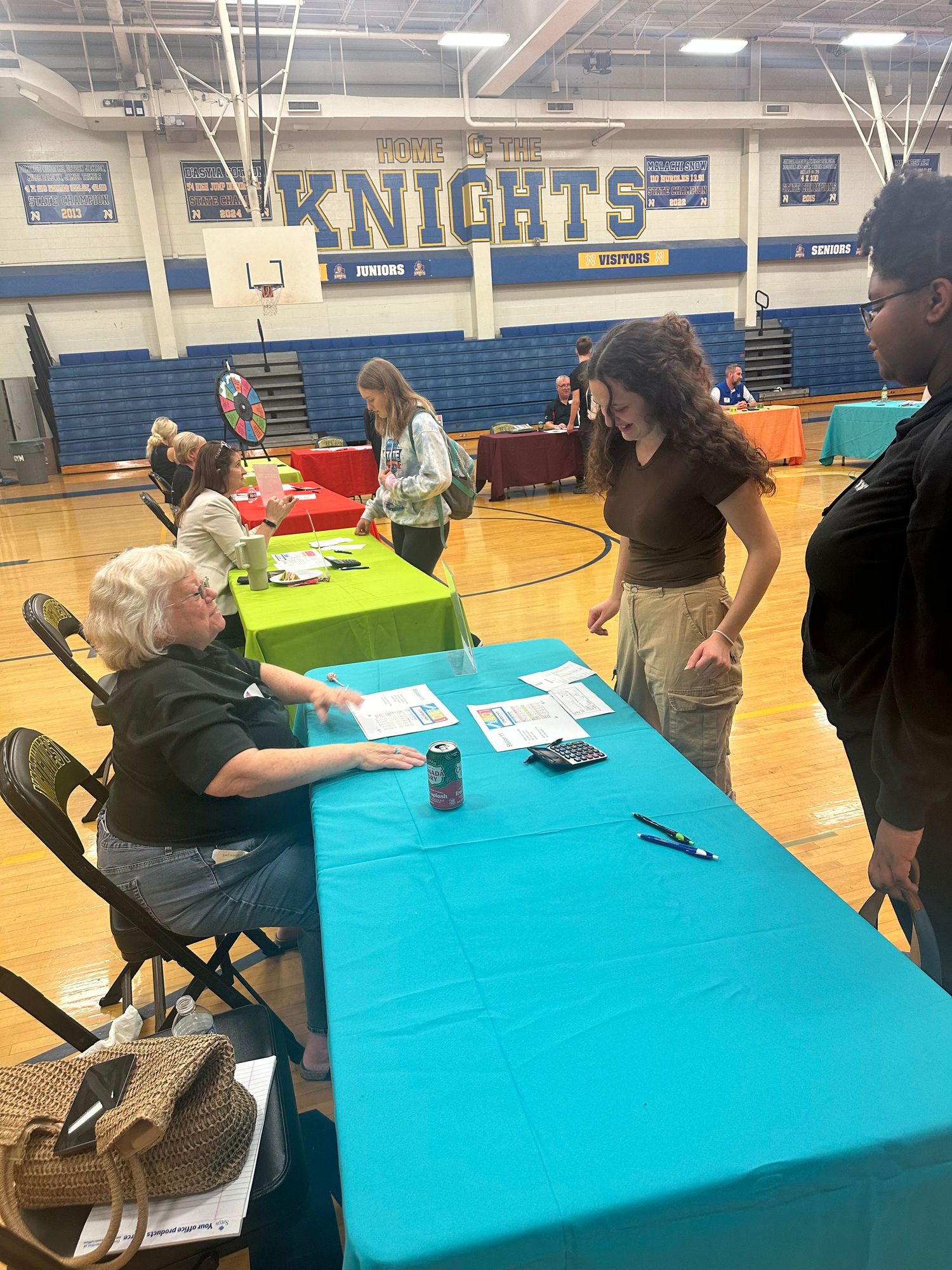 Students and staff interact at a Knights school gymnasium career fair with colorful tables on display.