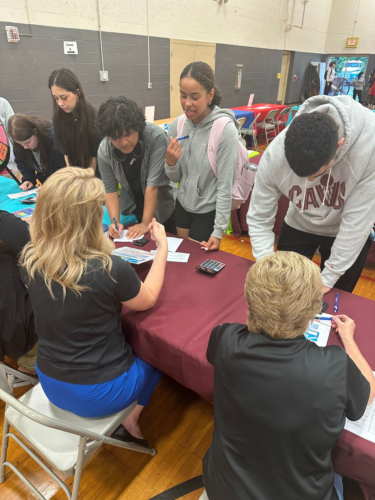 Students gather around a maroon table in a school gymnasium, reviewing documents with two seated adult advisors.