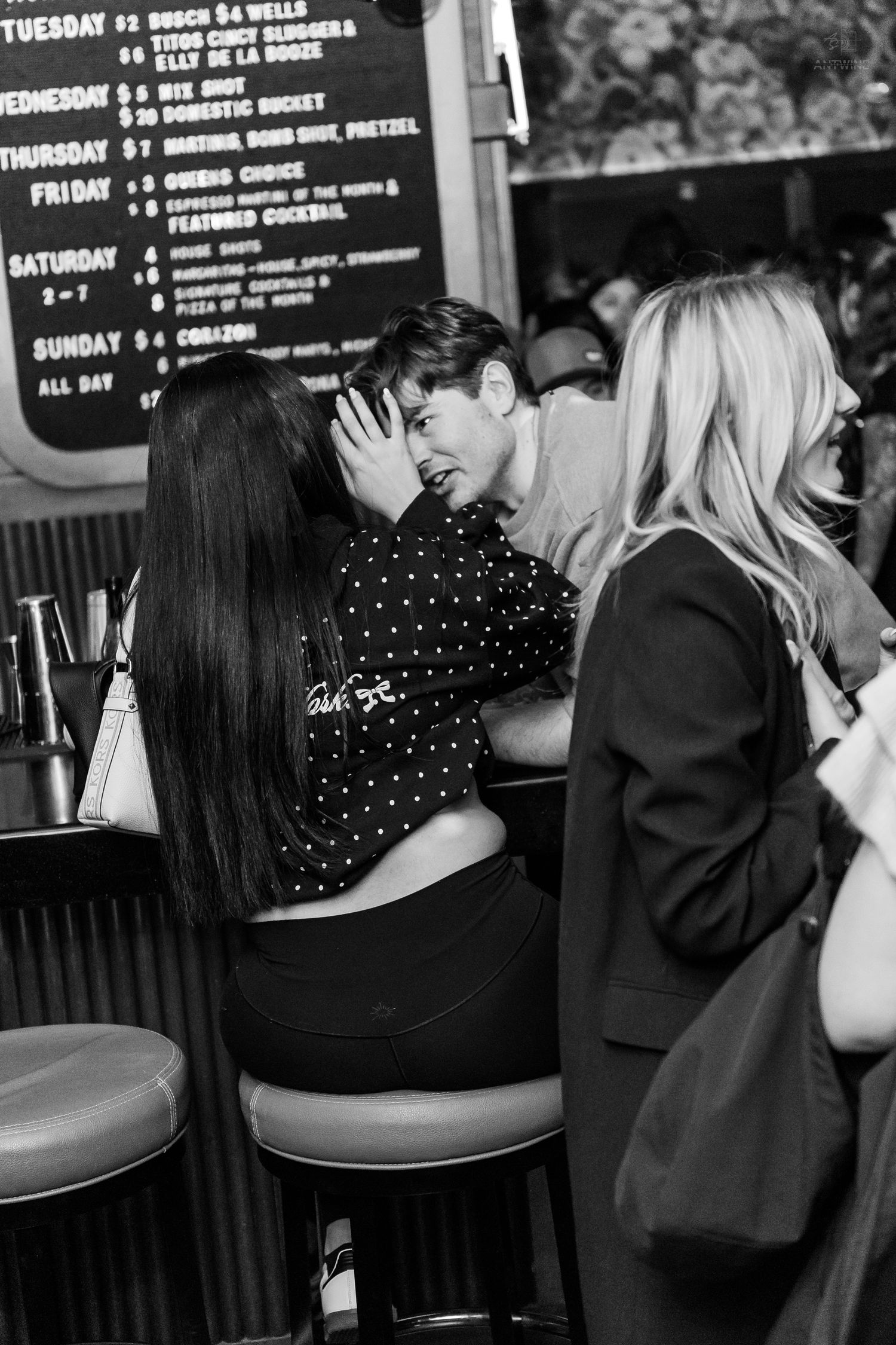 Young couple sharing a kiss at a bar while a blonde woman stands nearby in black and white.