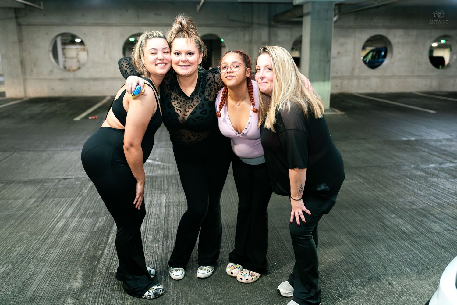 Four smiling women in black outfits pose together in an urban parking garage setting.