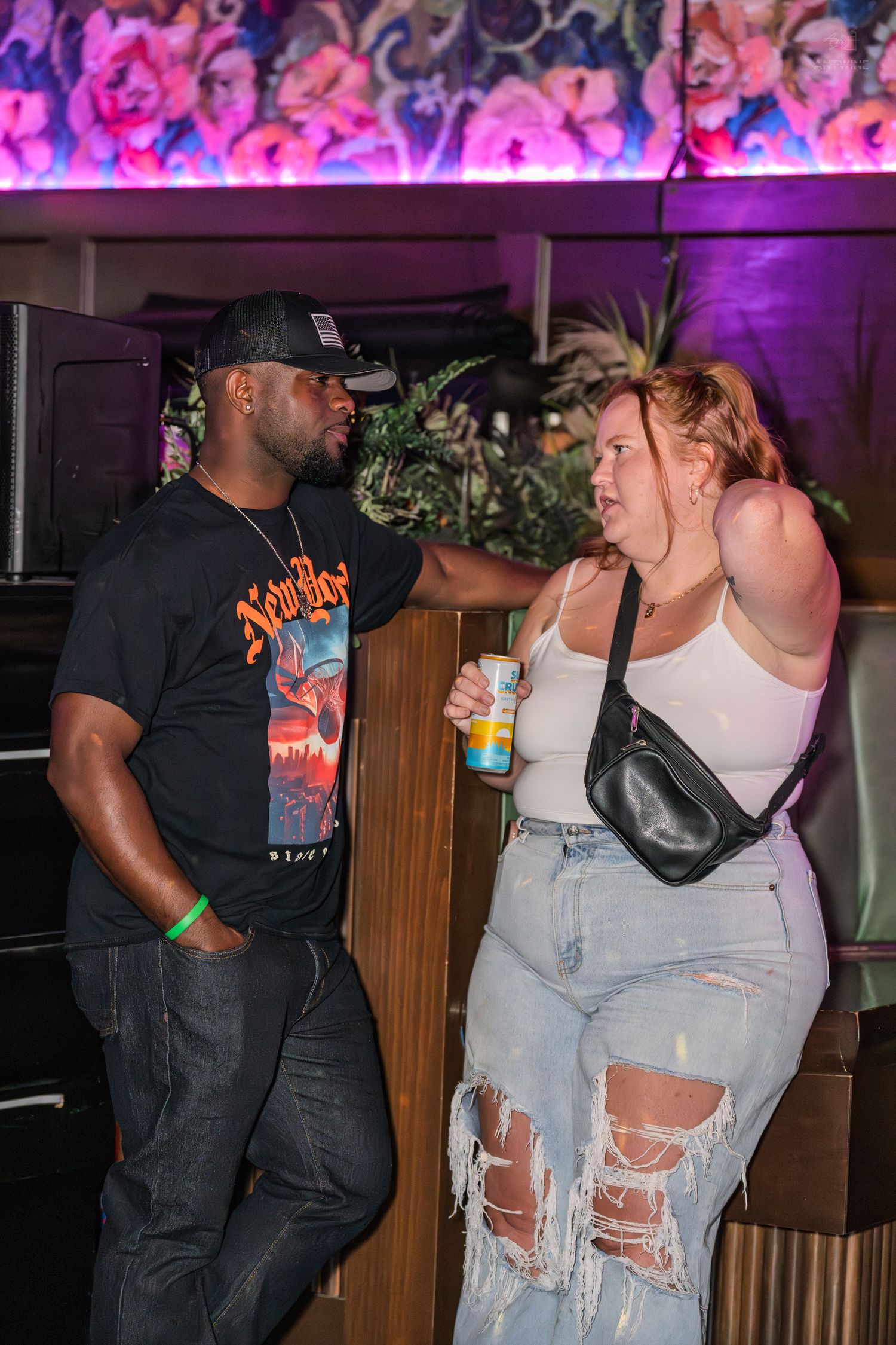 Man in black cap and graphic tee chats with woman holding drink in ripped jeans at nightclub.