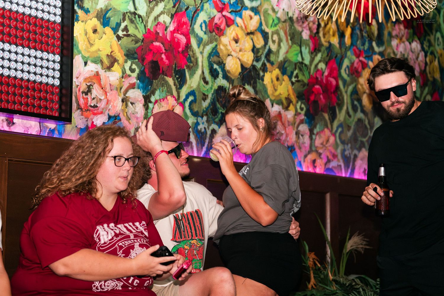 Group of people socializing at a bar with colorful floral artwork on the wall behind them.