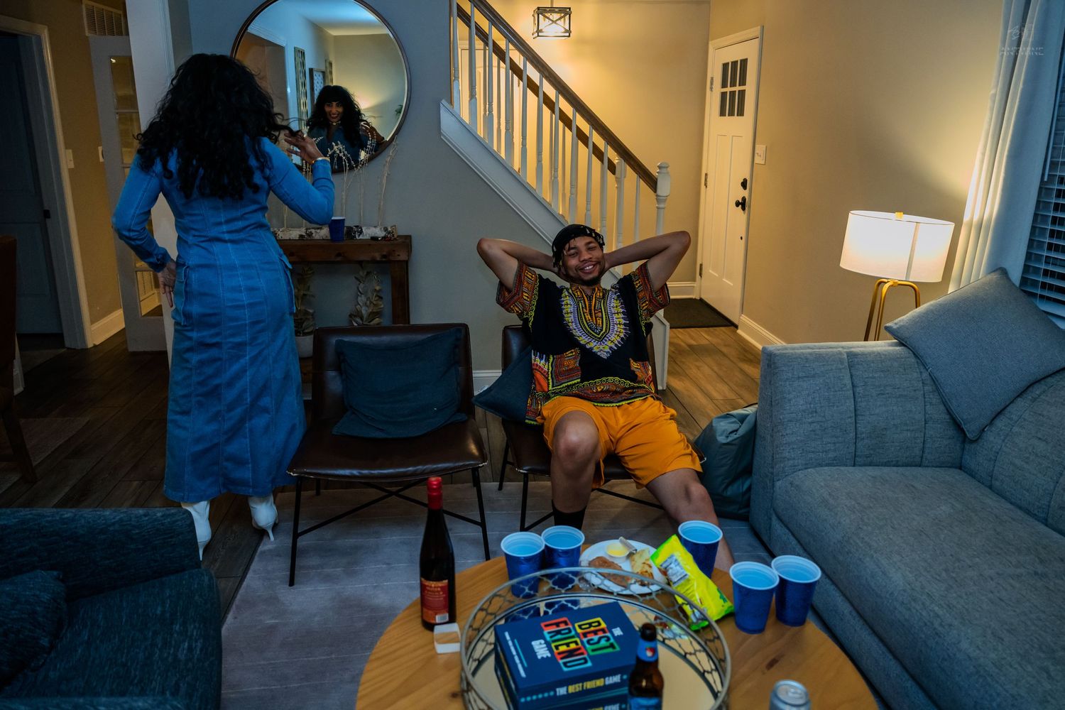 Friends gathering in living room with party supplies on coffee table, woman fixing hair near round mirror.