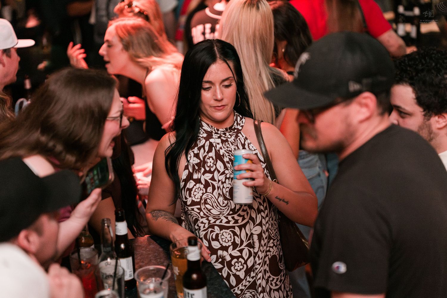 A dark-haired woman in a floral dress holds a drink can while standing in a crowded nightclub.