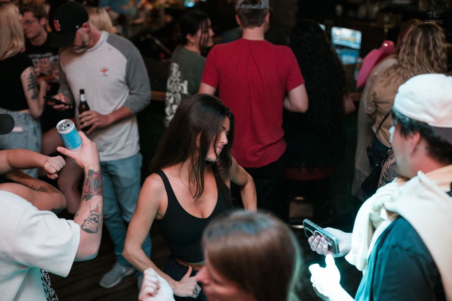 Woman in black tank top laughing while people hold drinks at a lively bar party.
