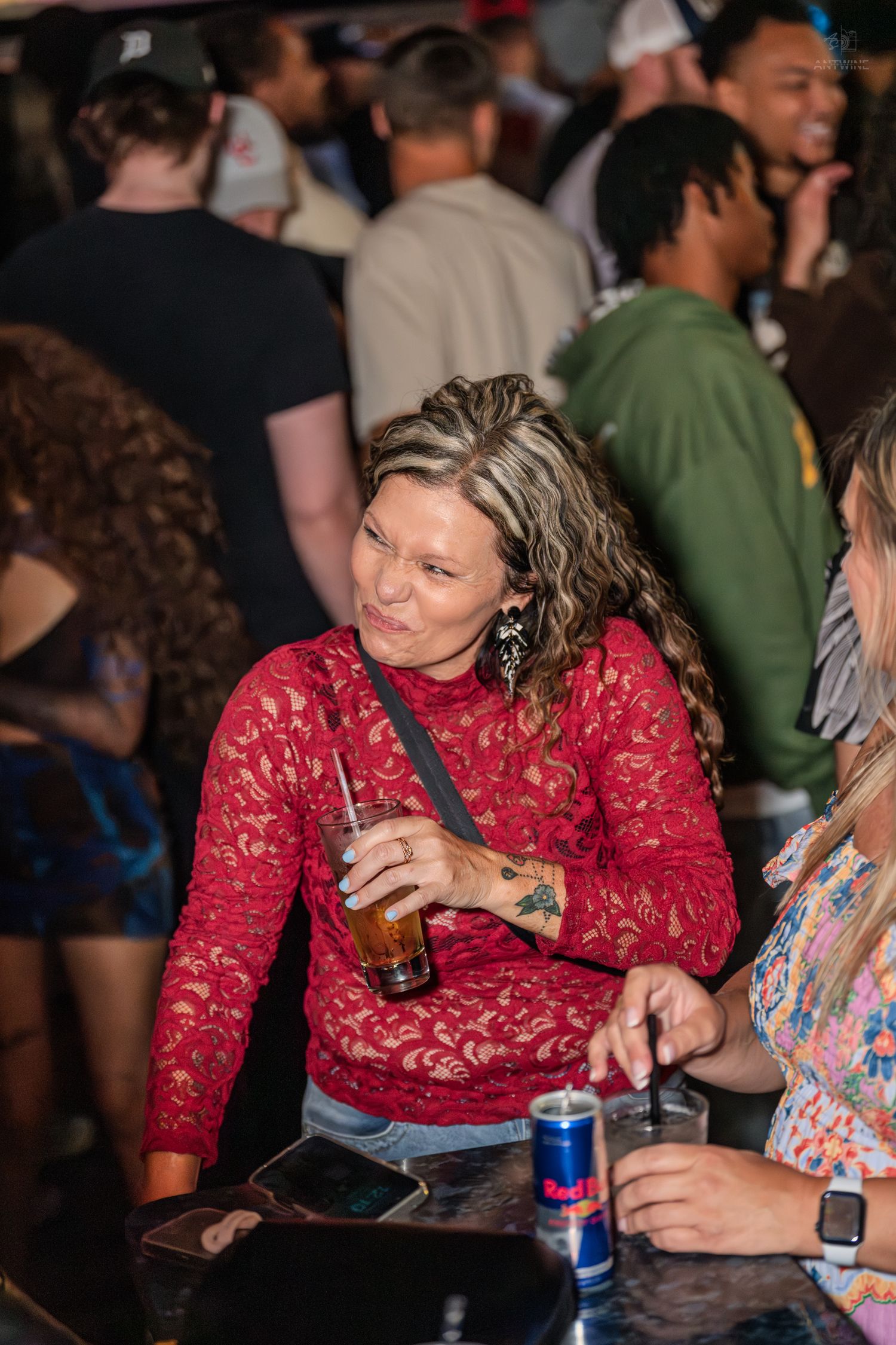 Woman in red lace top holding a drink at a crowded bar.