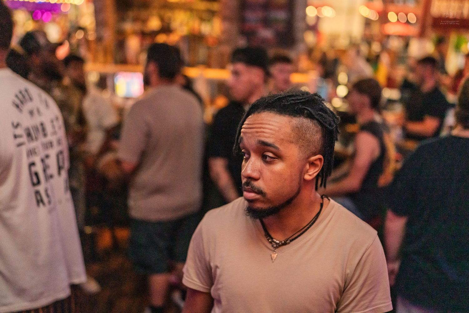 Young Black man with braided hair in beige t-shirt stands in a busy bar crowd.