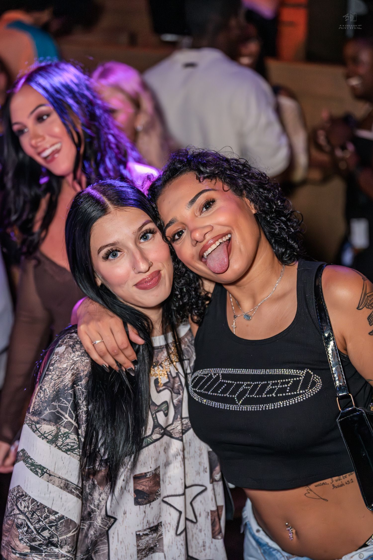 Two young women pose together at a nightclub, one sticking out her tongue, with purple lights and crowd behind them.