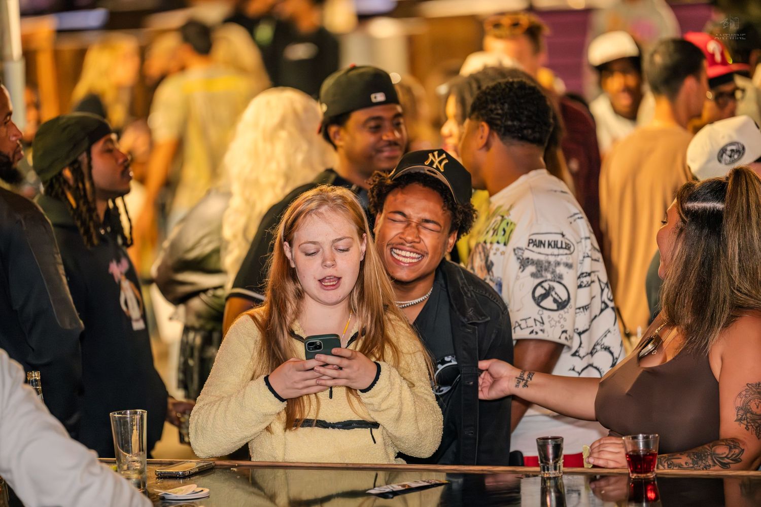 Two young people smiling and looking at a phone together at a busy nightlife event.
