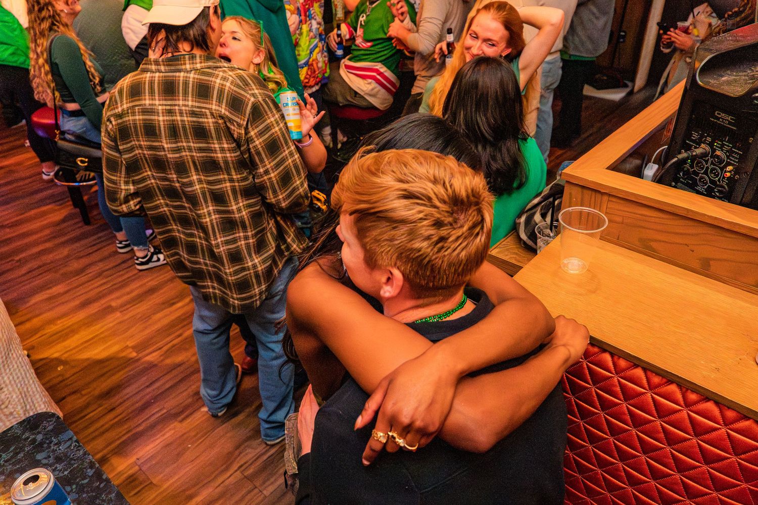 Two people embracing at a crowded indoor bar event with wooden floors and red booth seating.