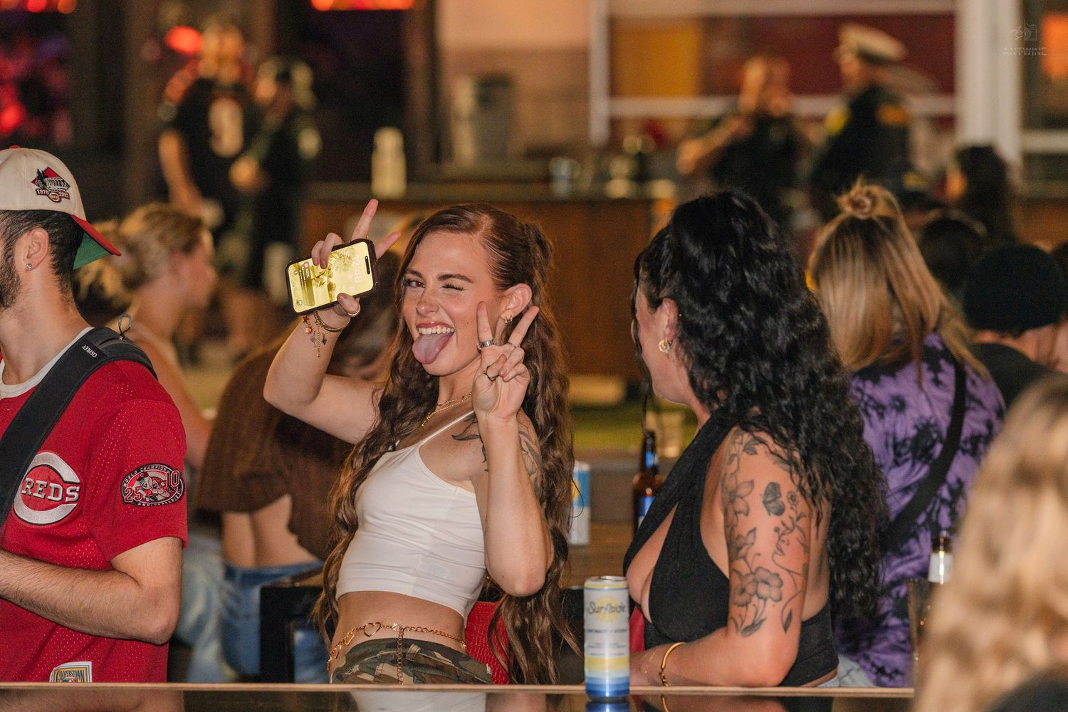 Woman taking a selfie and flashing a peace sign with a tattooed friend at a lively bar event.