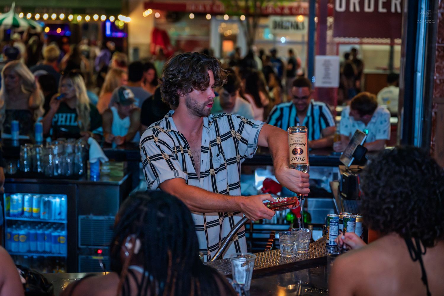 A male bartender in a checkered shirt pours a bottle of liquor at a crowded bar.
