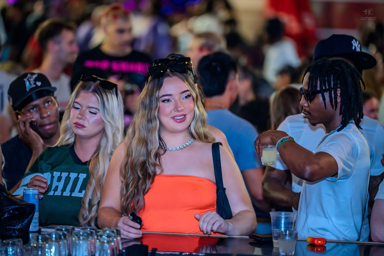 Smiling woman in orange top poses at a bar in a busy nightclub surrounded by friends.