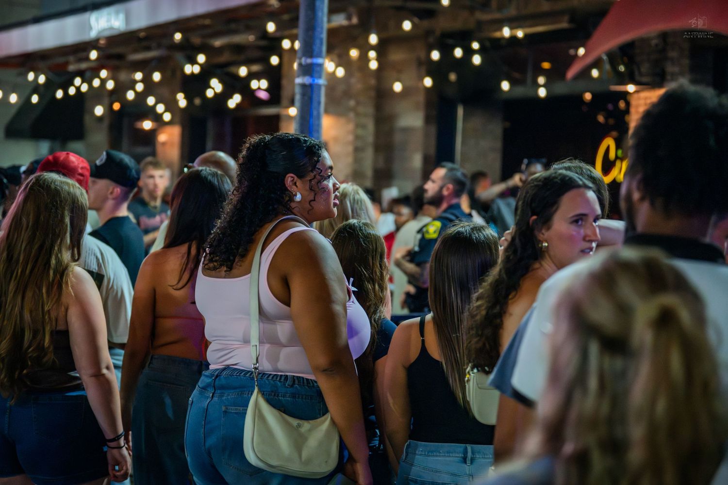 A crowd of people socializing at a lively outdoor nighttime event with string lights in the background.