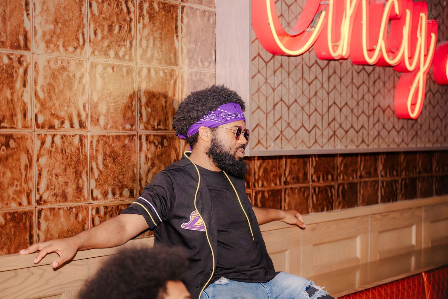 Stylish man in purple bandana and sunglasses posing near a Cincy neon sign.