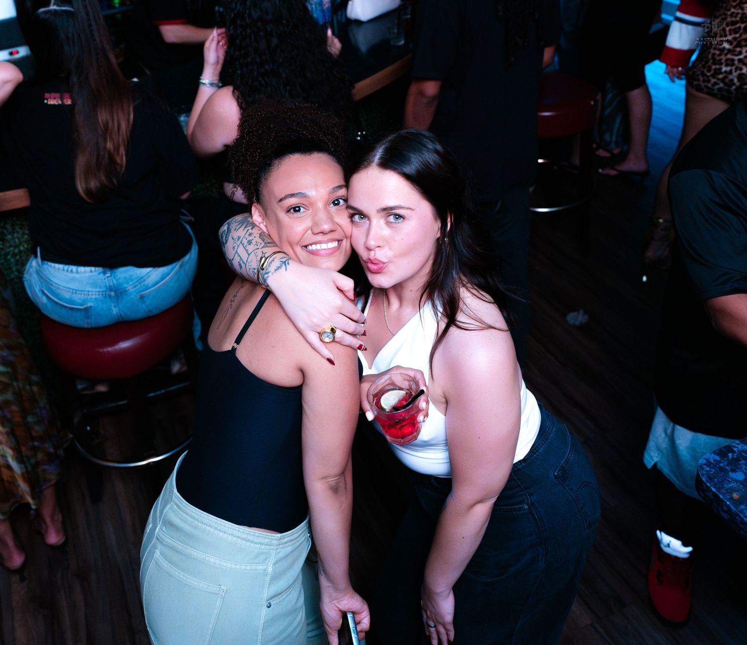 Two friends hugging and posing with drinks at a nightclub party.