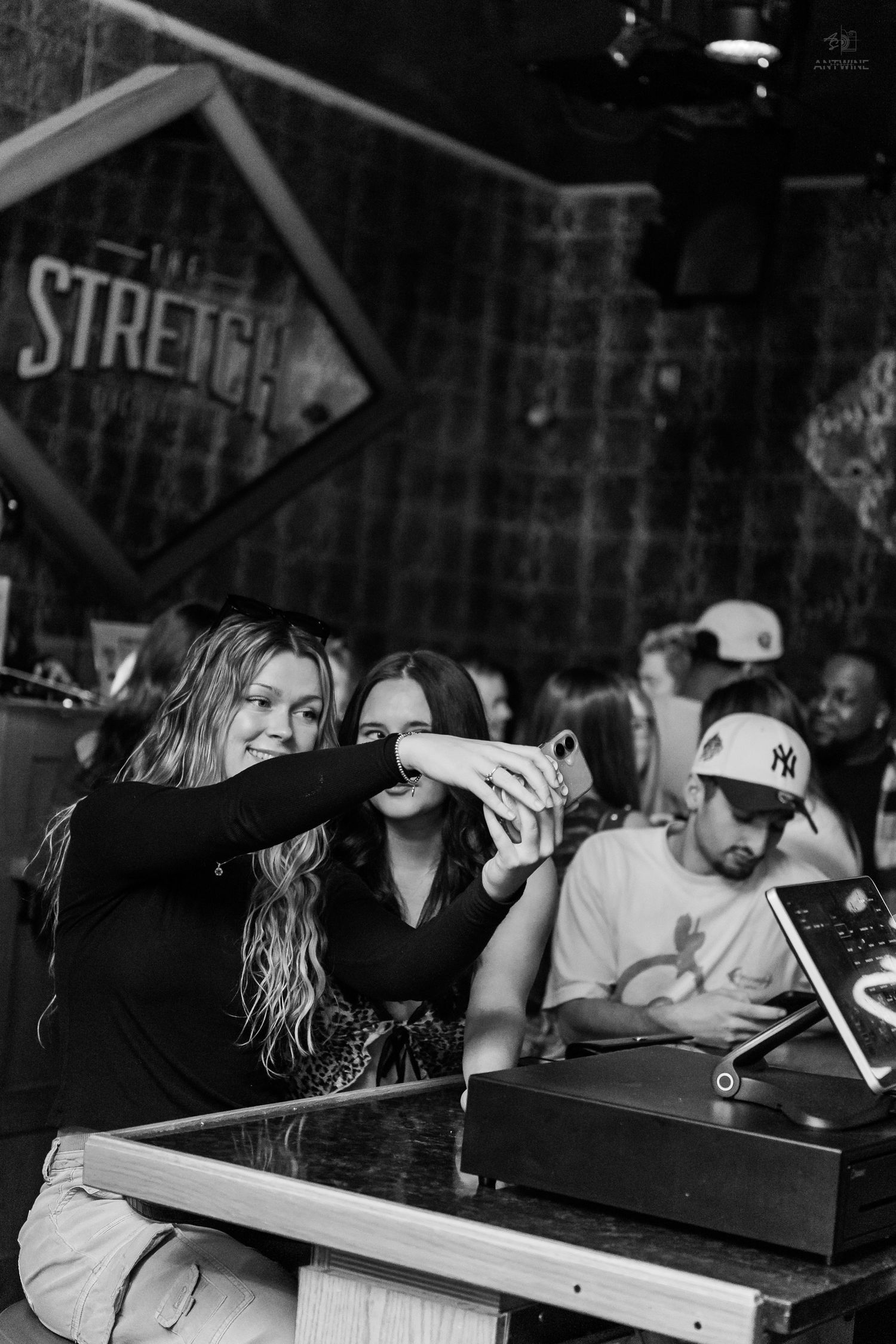Two women take a selfie at The Stretch bar while a man in an NY Yankees cap looks on.