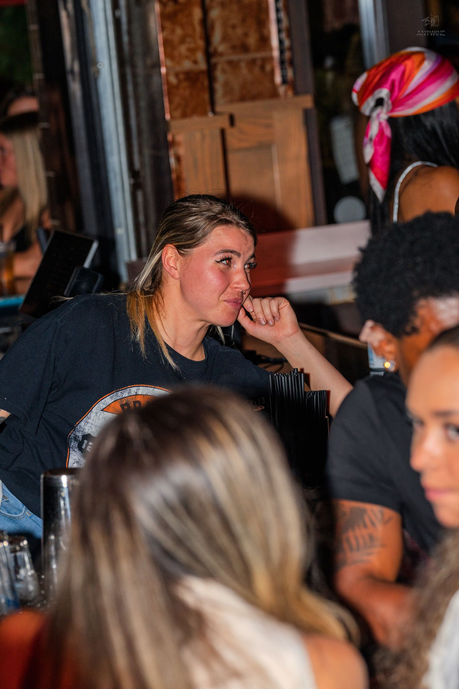 Blonde woman in black shirt rests chin on hand while seated at a busy social gathering.