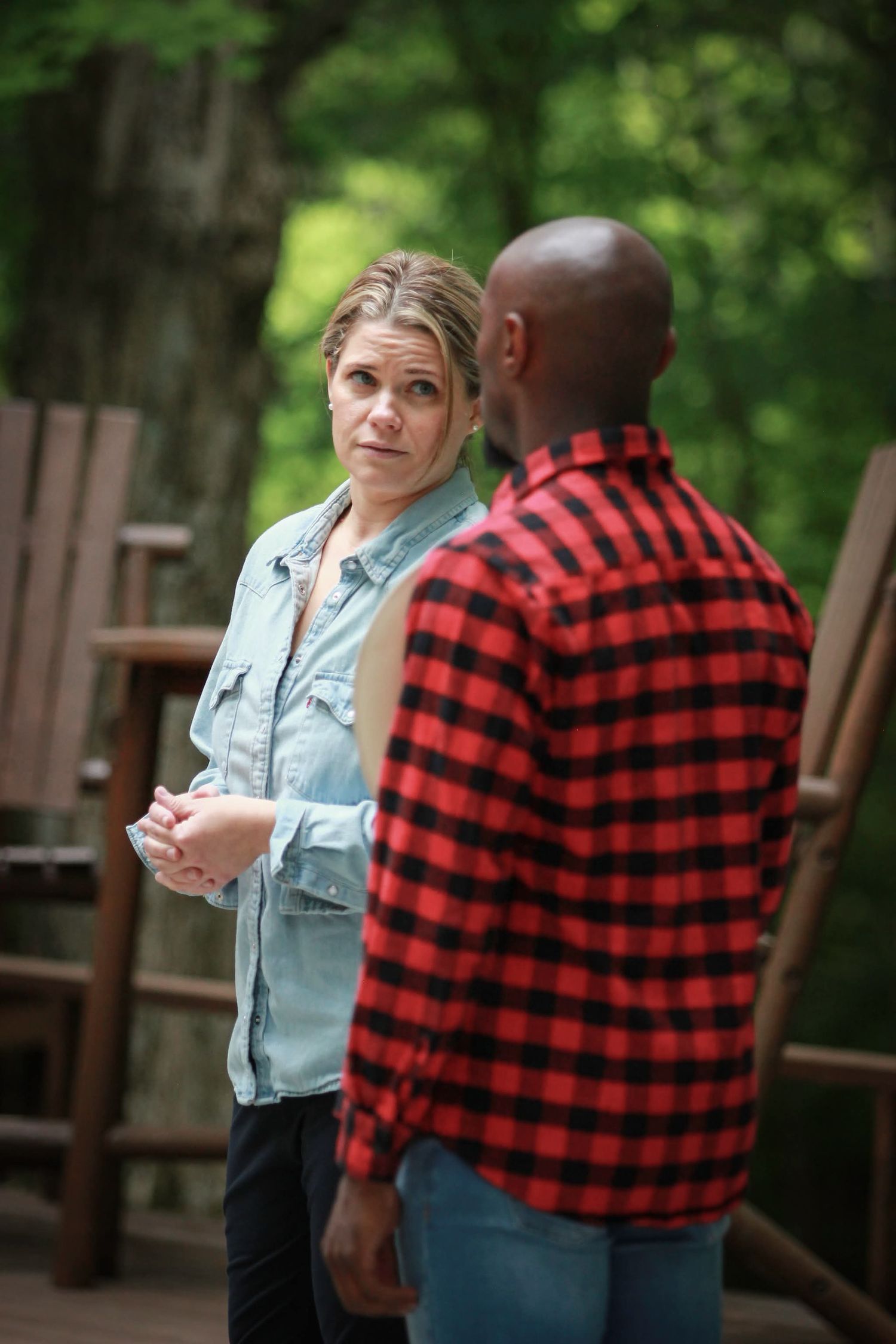 A woman in a denim shirt and a man in a red plaid shirt having a conversation on a wooded deck.