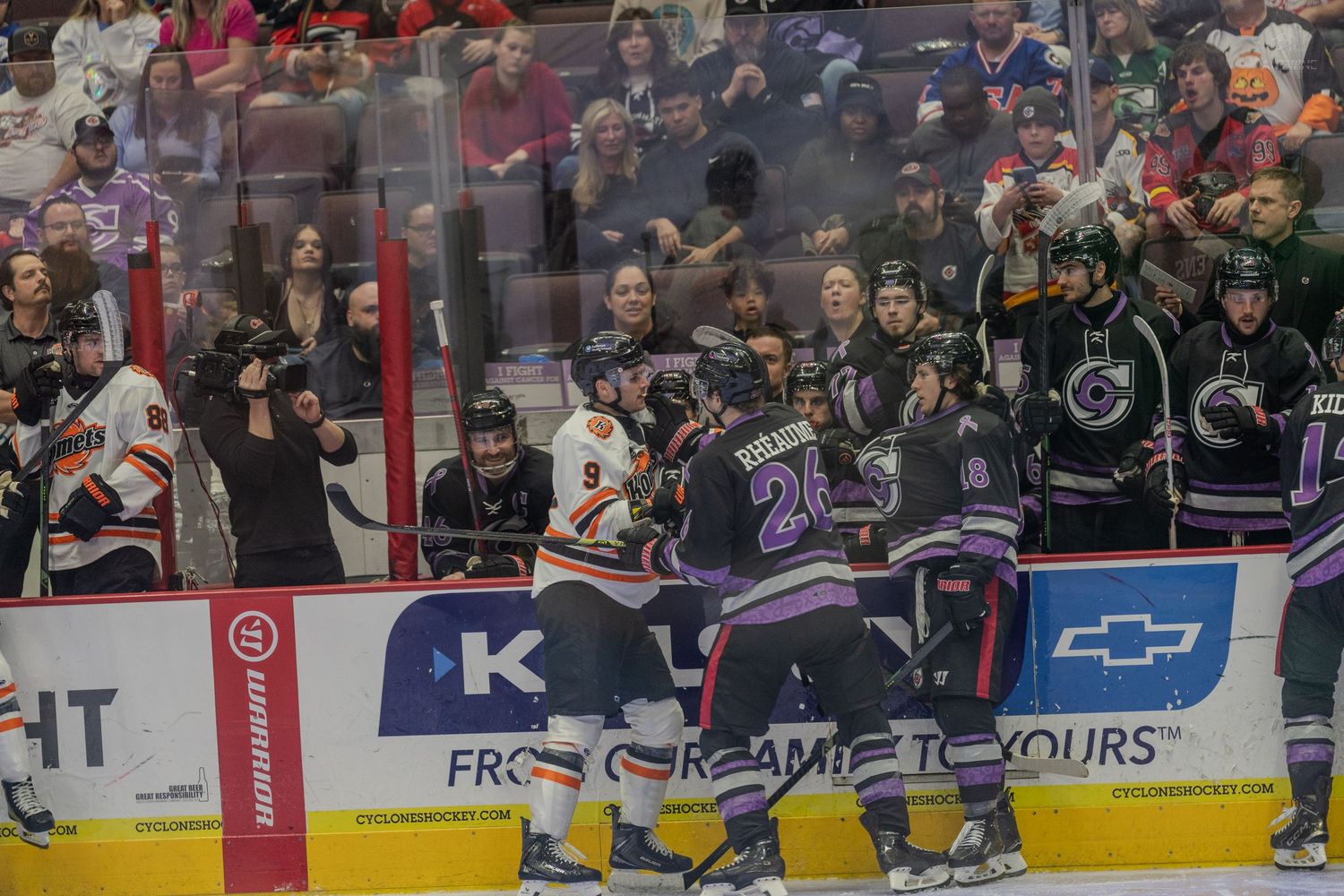 Hockey players in purple and orange jerseys engage in physical scrum along boards during intense game play.