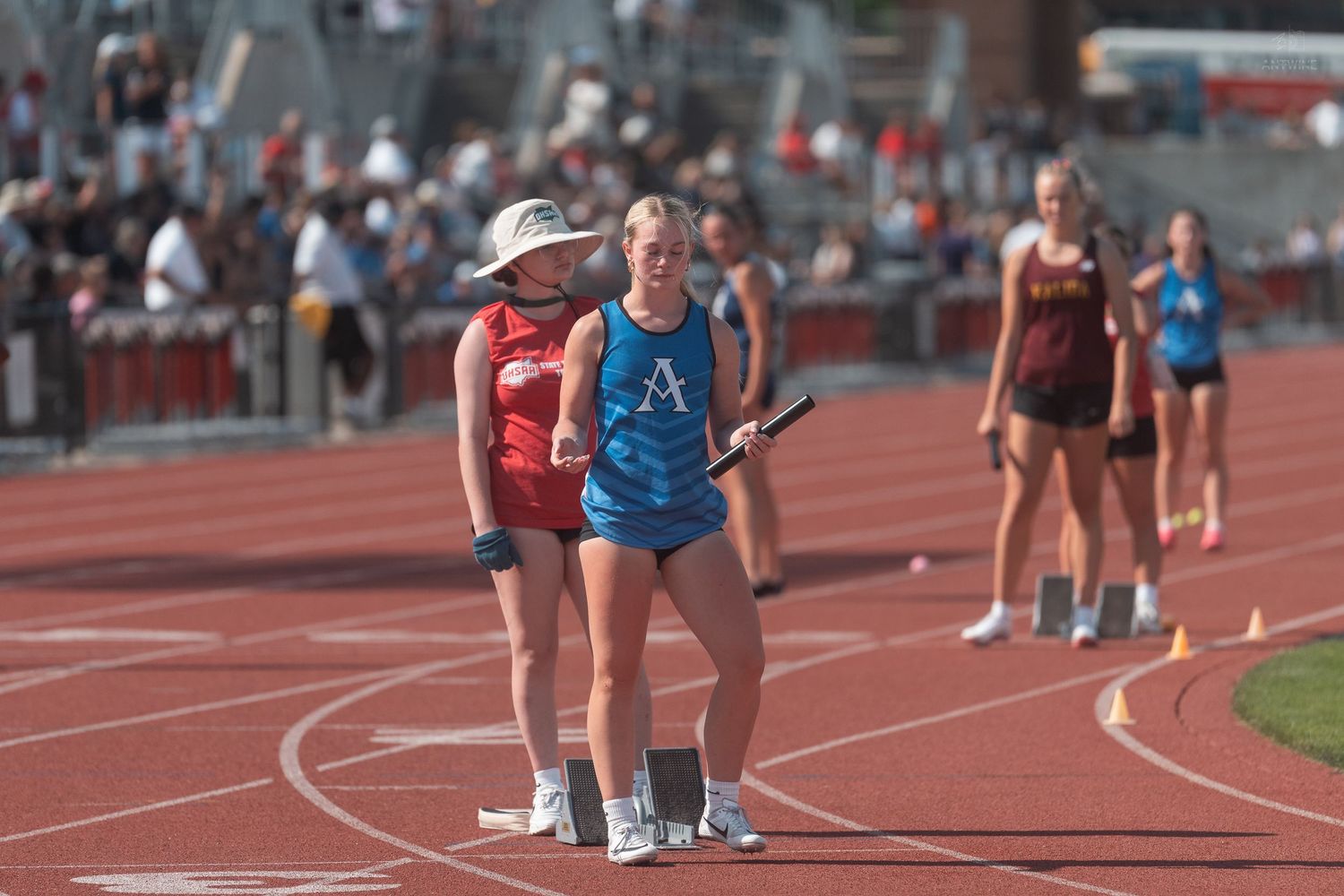 Female athlete in blue uniform holds relay baton beside coach in sun hat on a red track.