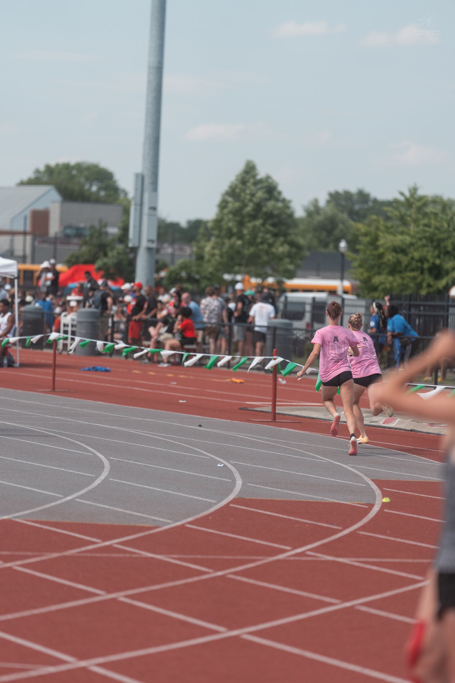 Two female runners in pink shirts race on a red track during an outdoor track and field competition.