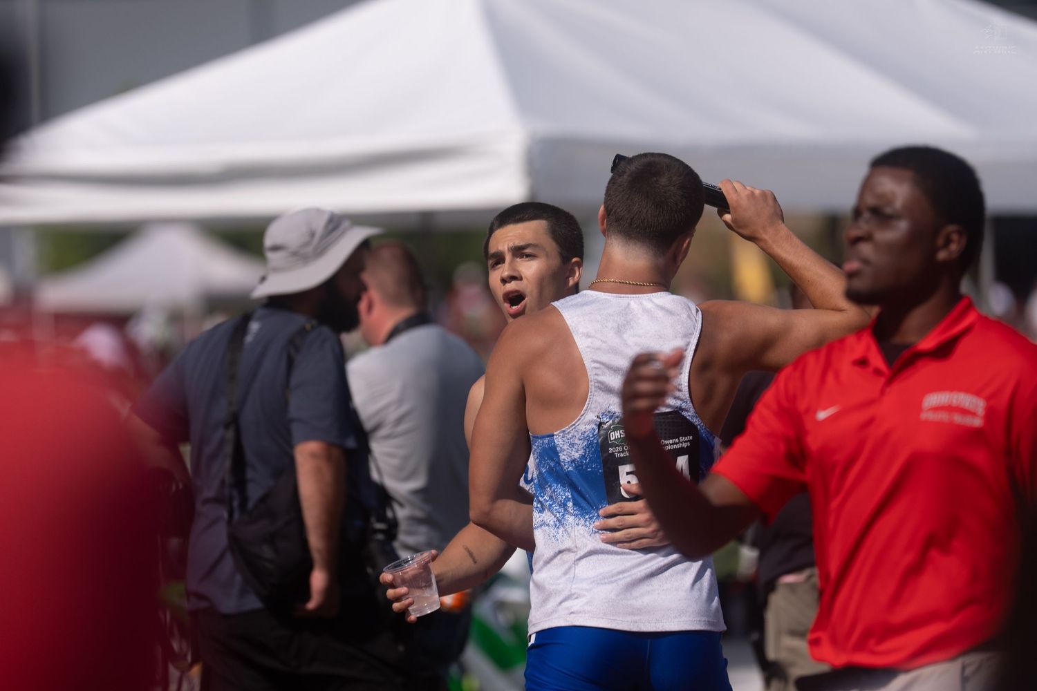 Athletes embrace and celebrate at an outdoor track and field competition under white tents on a sunny day.