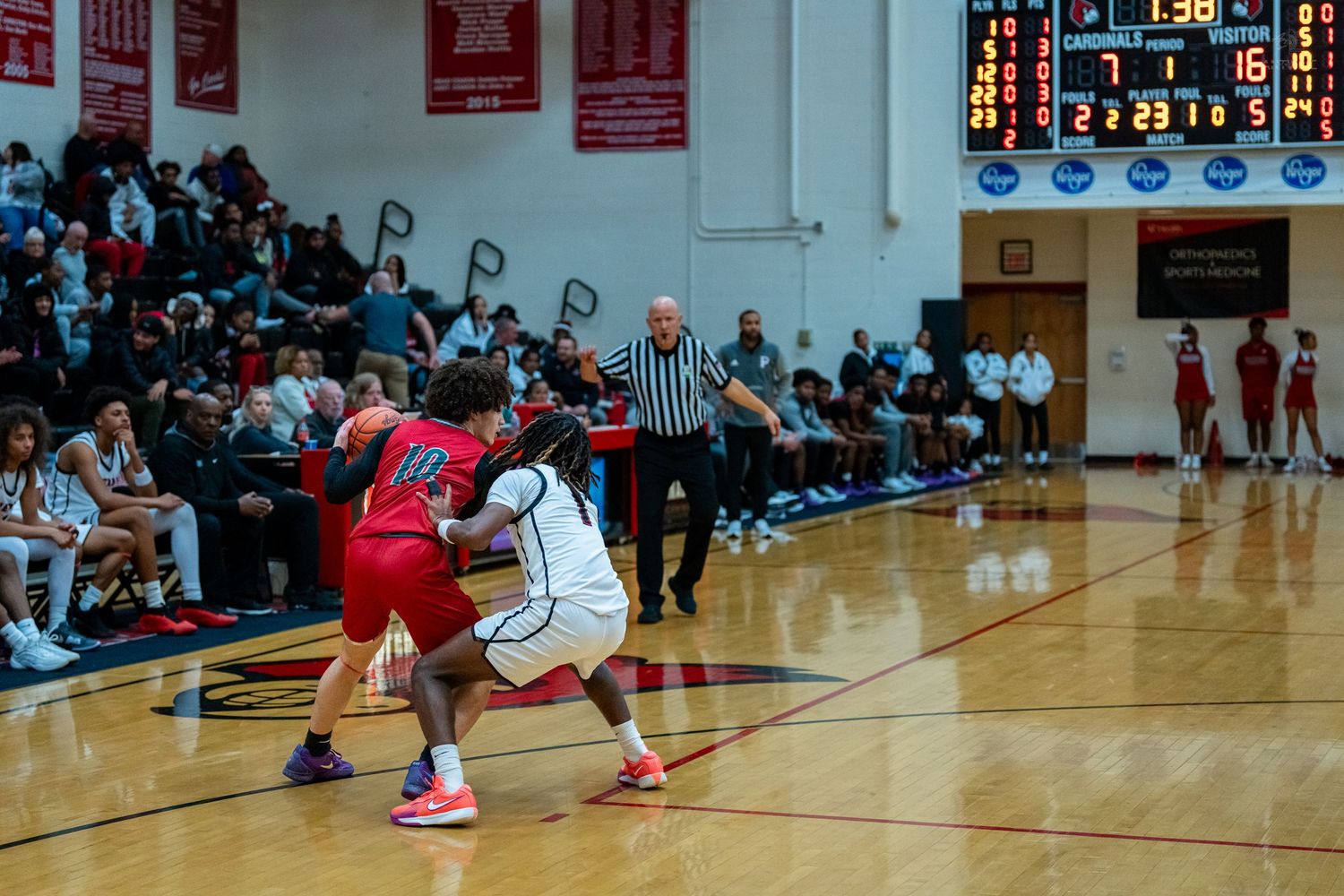 High school basketball game in progress with players defending near the sideline, scoreboard visible in background.