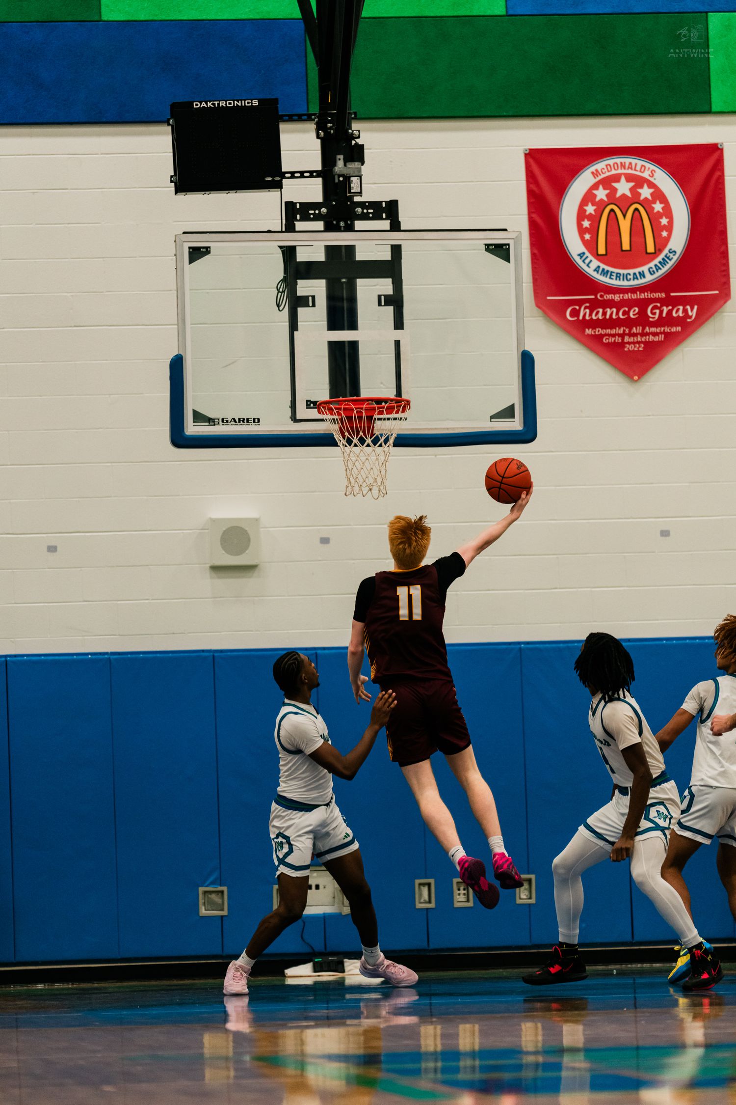 Player #11 in dark jersey leaps for a layup during a high school basketball game in a blue and green gymnasium.