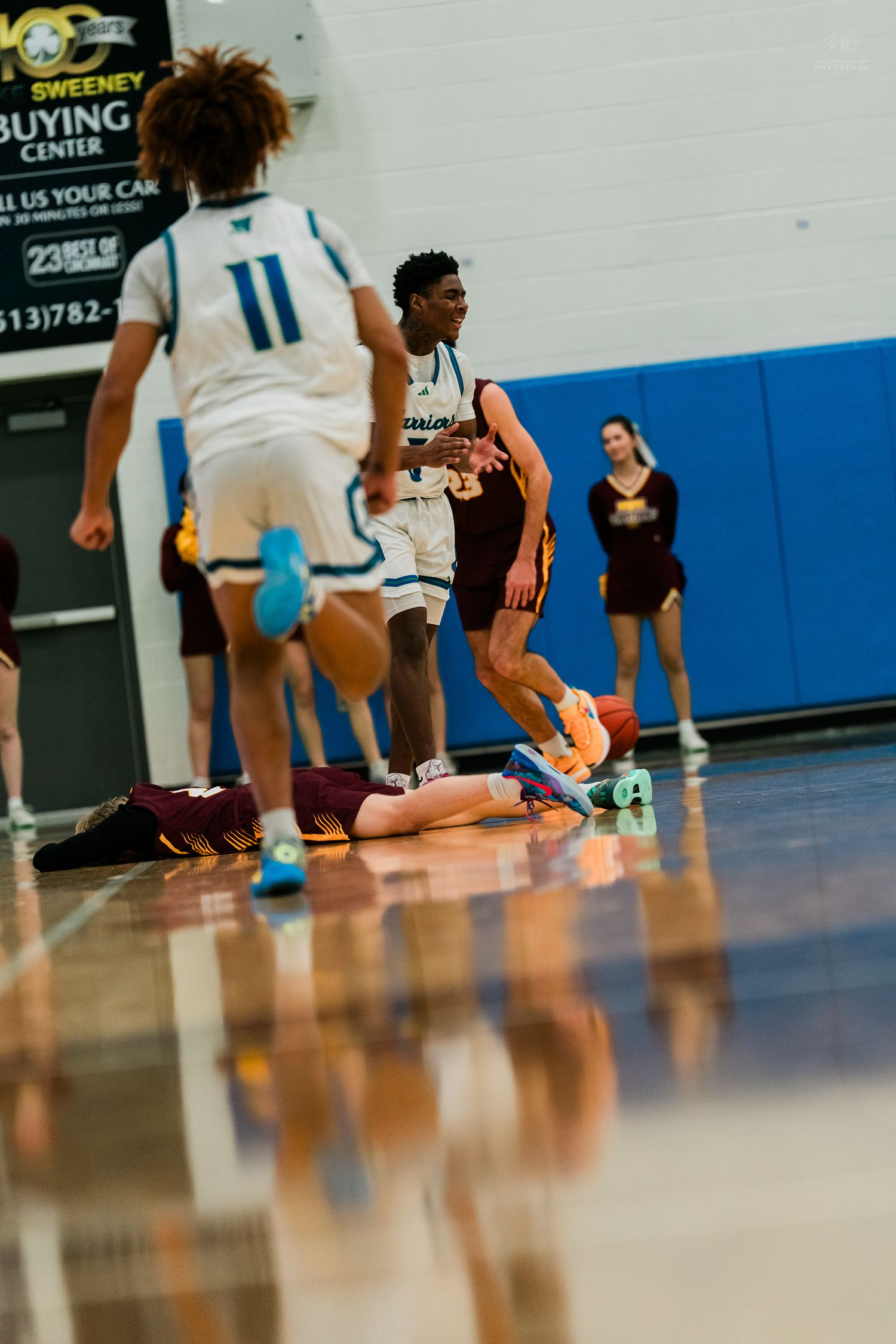 A basketball player in white Warriors uniform stands over a fallen opponent during an intense high school basketball game.