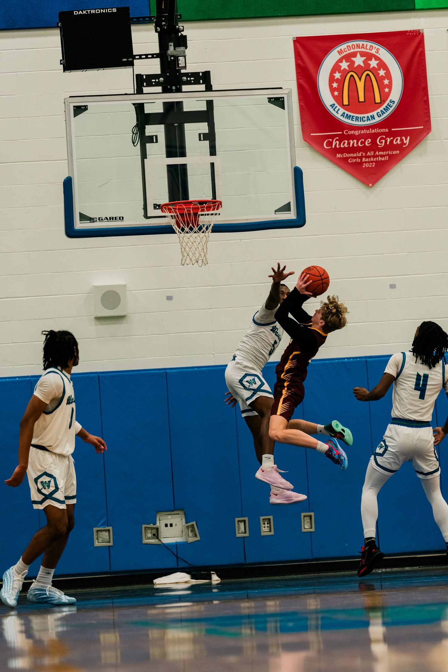 A basketball player attempts a layup while being defended during a high school game in a blue and white gymnasium.