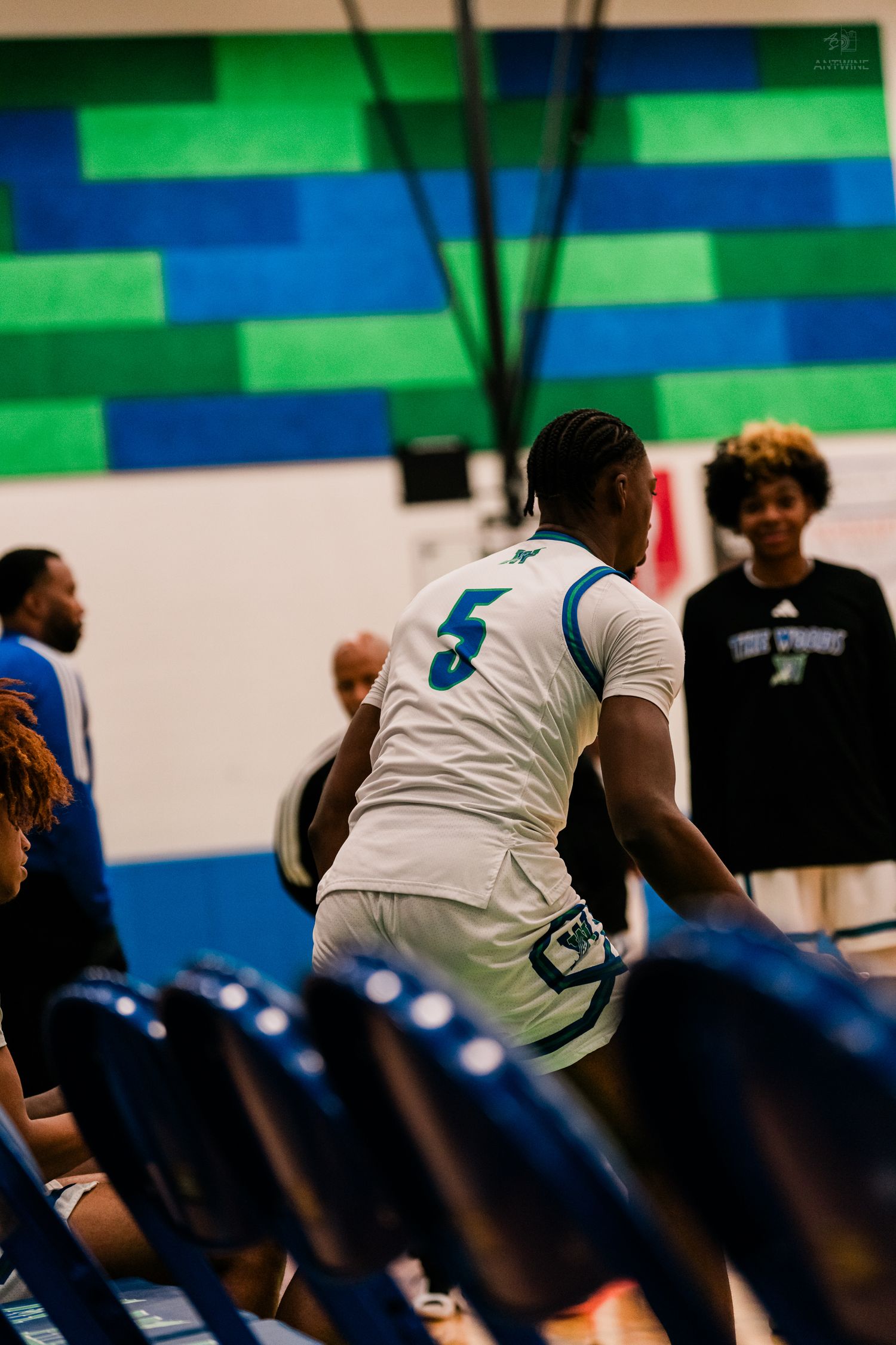 High school basketball player #5 in white uniform walks toward bench area in colorful gym.