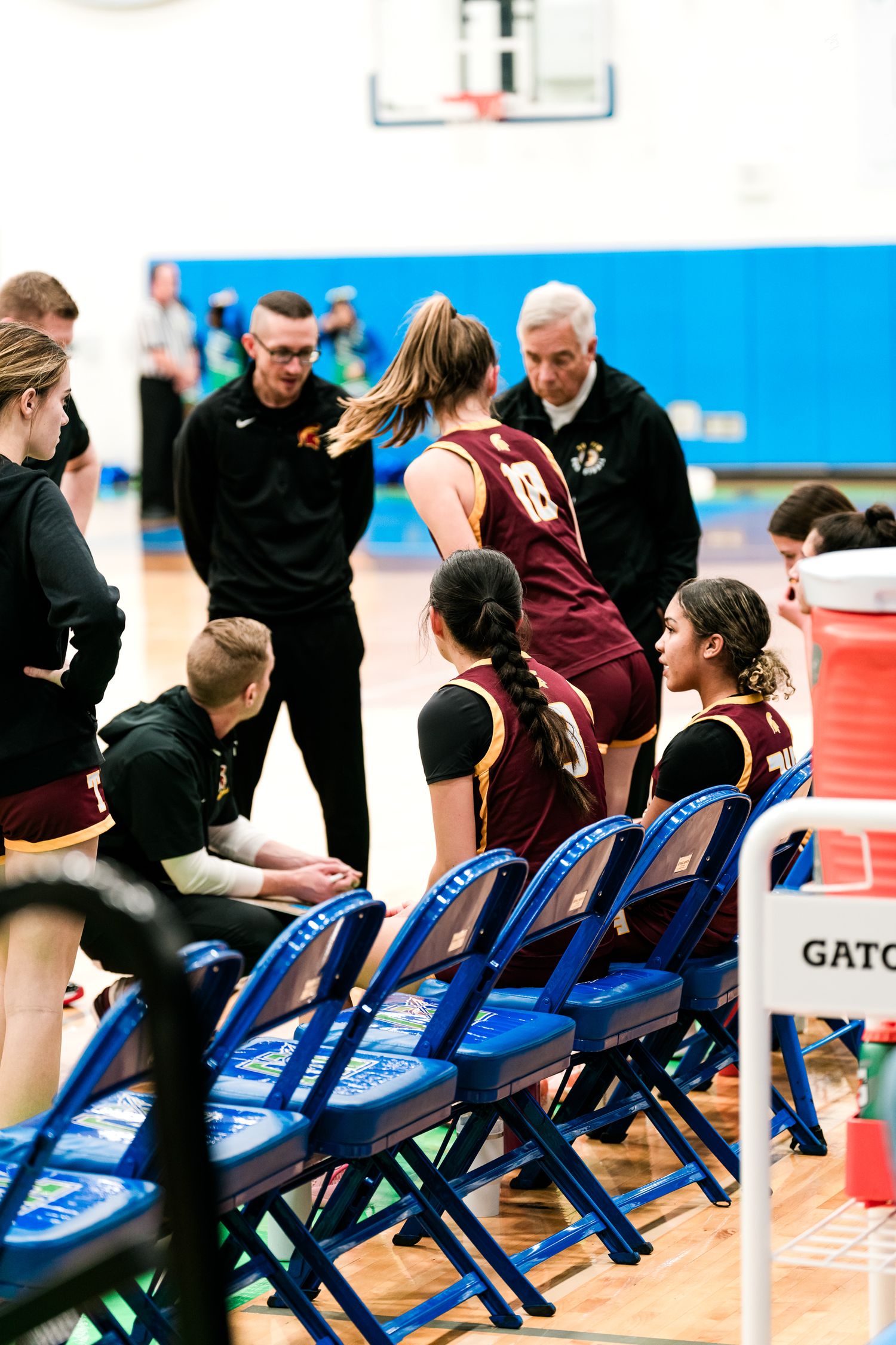 Basketball coaches in black uniforms huddle with young female players in maroon jerseys during a timeout on the sideline.